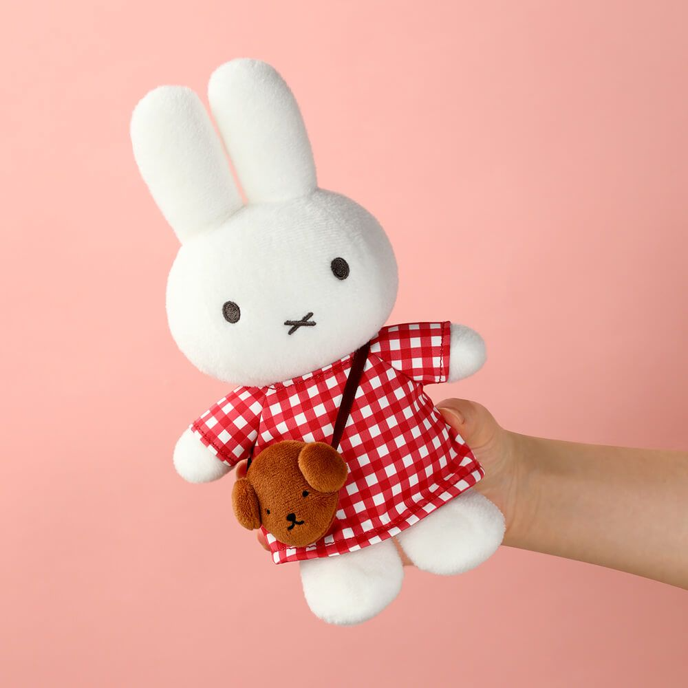 White plush rabbit toy in a red checkered dress held against a pink background
