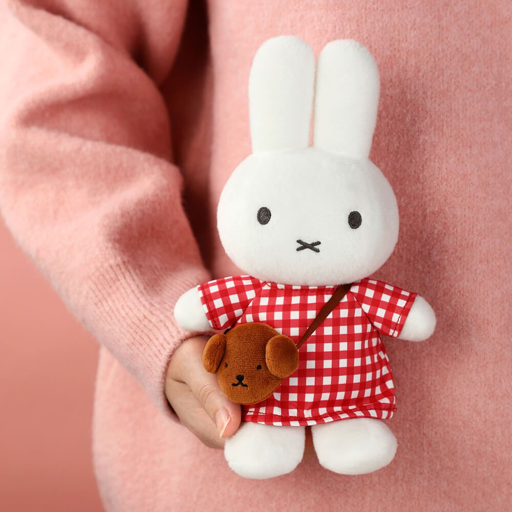 Plush toy rabbit in a red checkered dress held by a hand against a pink background