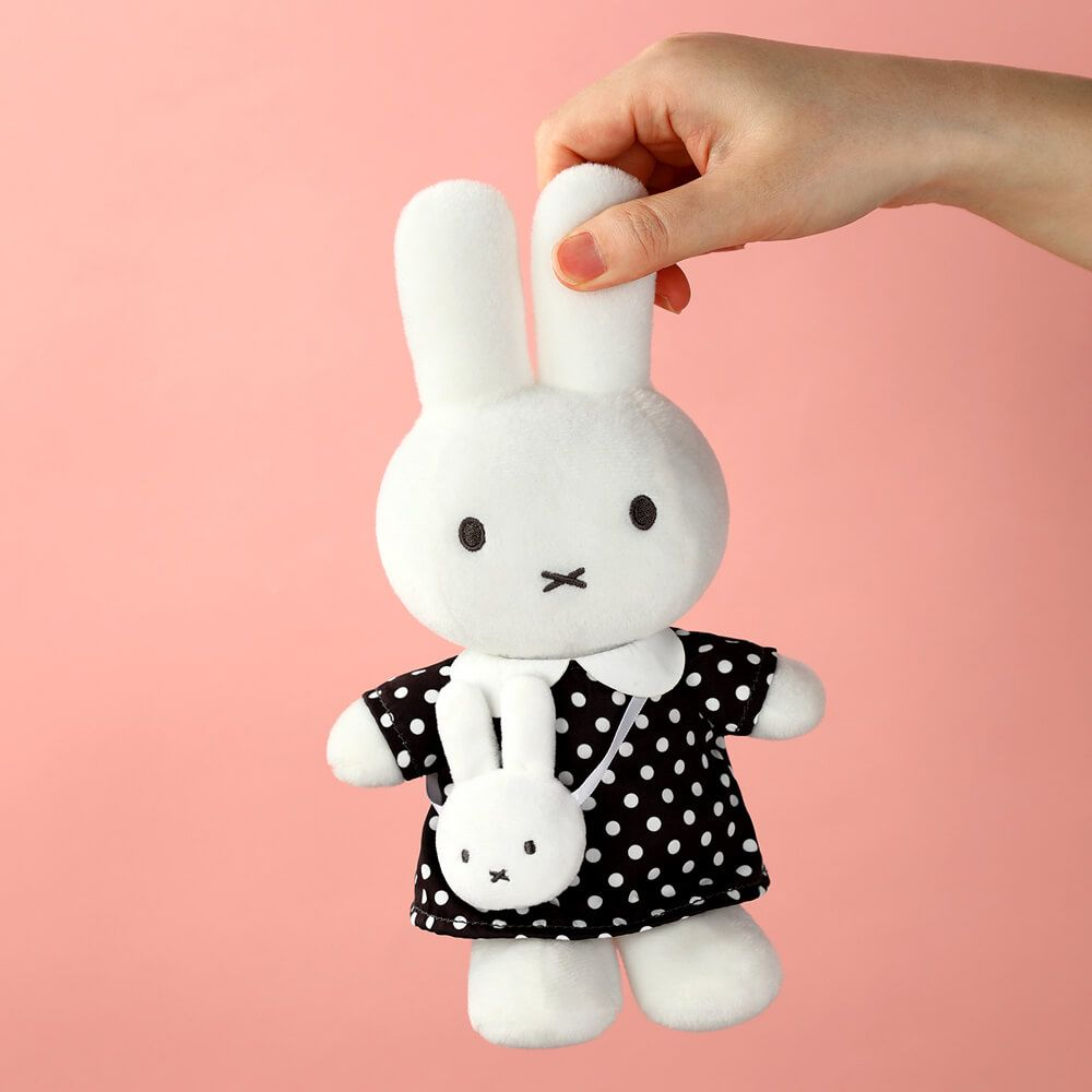 A white plush bunny doll wearing a black dress with white polka dots, held by a person's hand against a solid pink background.