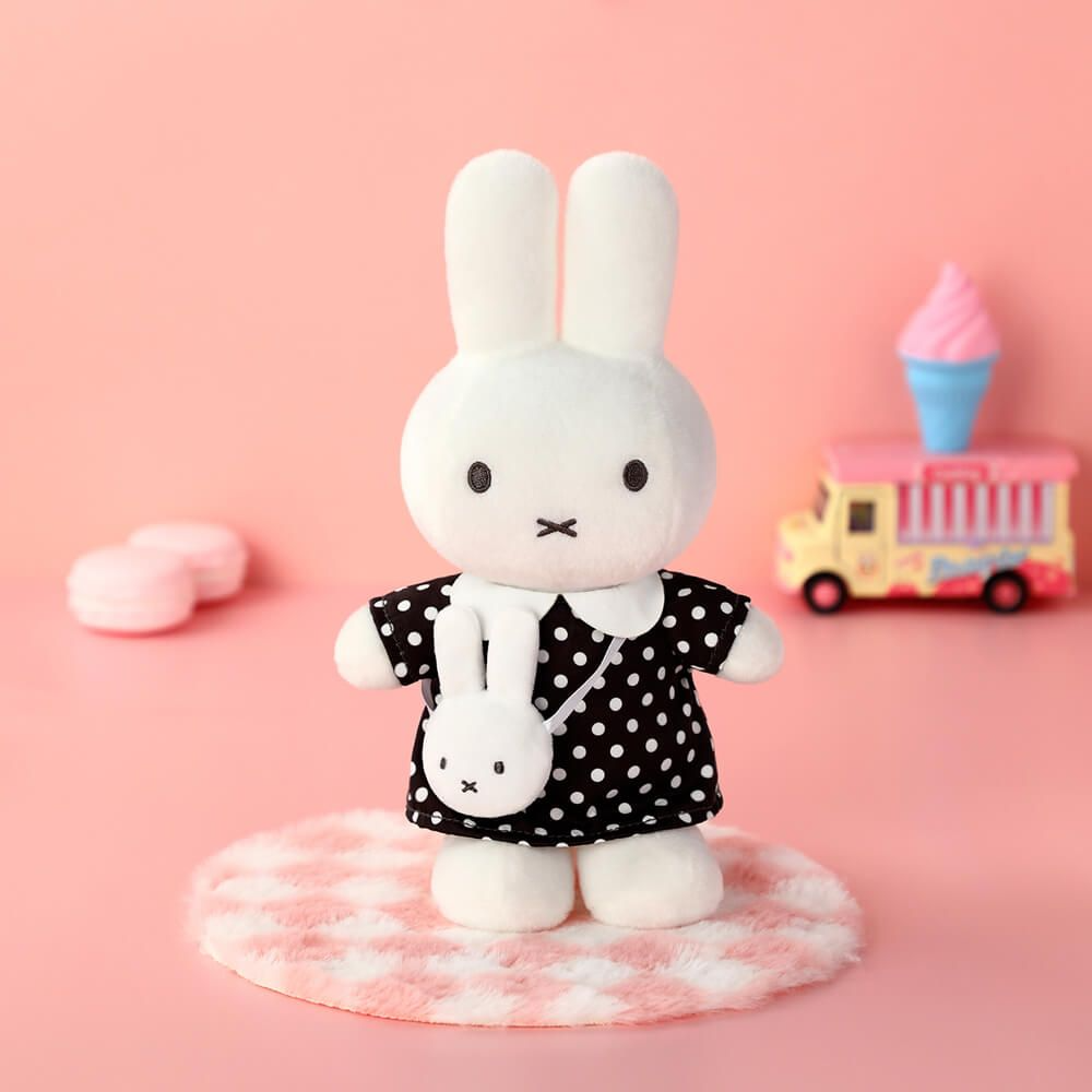 A white plush bunny doll wearing a black dress with white polka dots and a white collar, standing on a pink-and-white checkered rug against a soft pink background. A small matching bunny crossbody bag is hanging from the neck, with a toy ice cream truck and pink accessories blurred in the background.