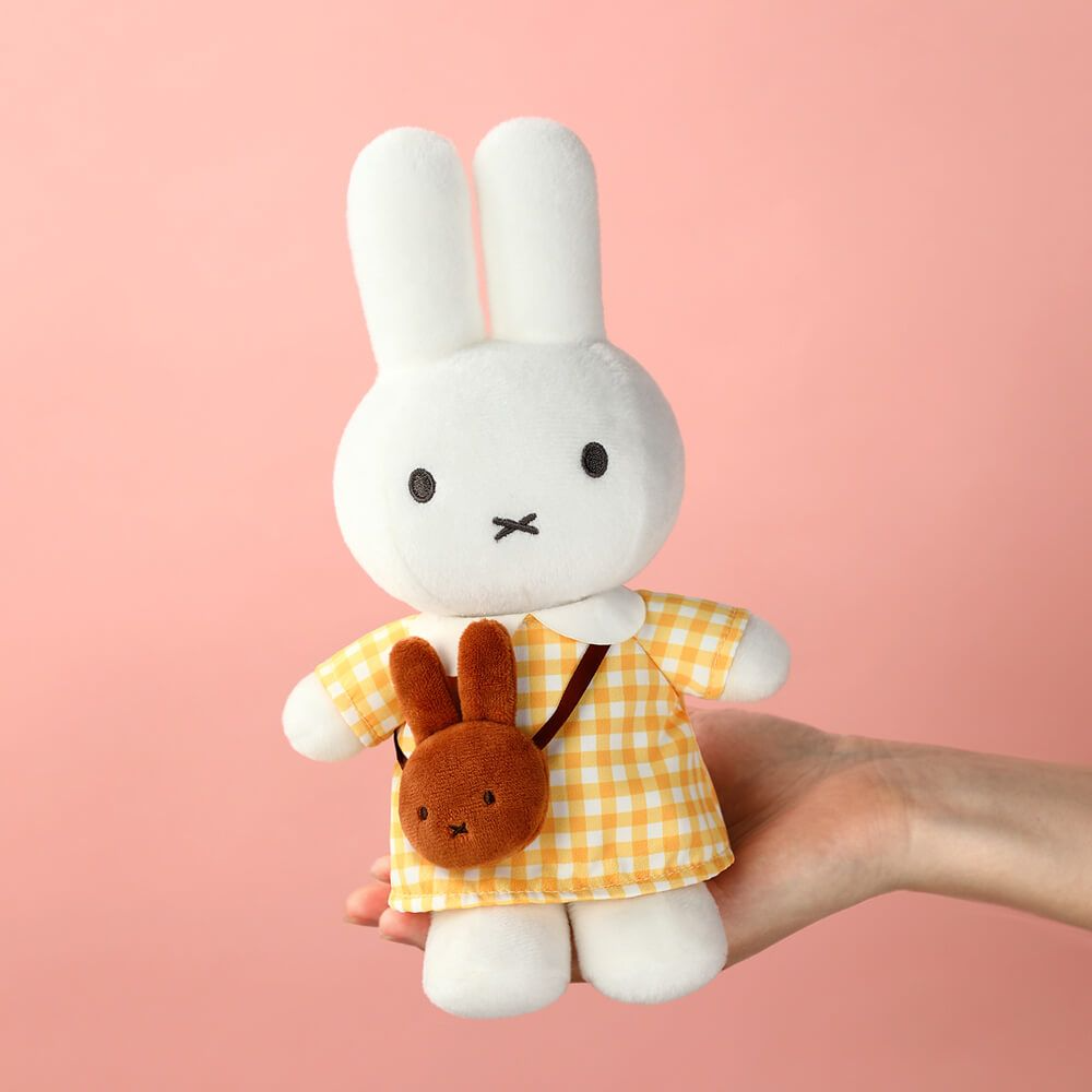 A white plush bunny doll wearing a yellow-and-white gingham dress and carrying a small brown bunny-shaped bag, held in a person's hand against a soft pink background.