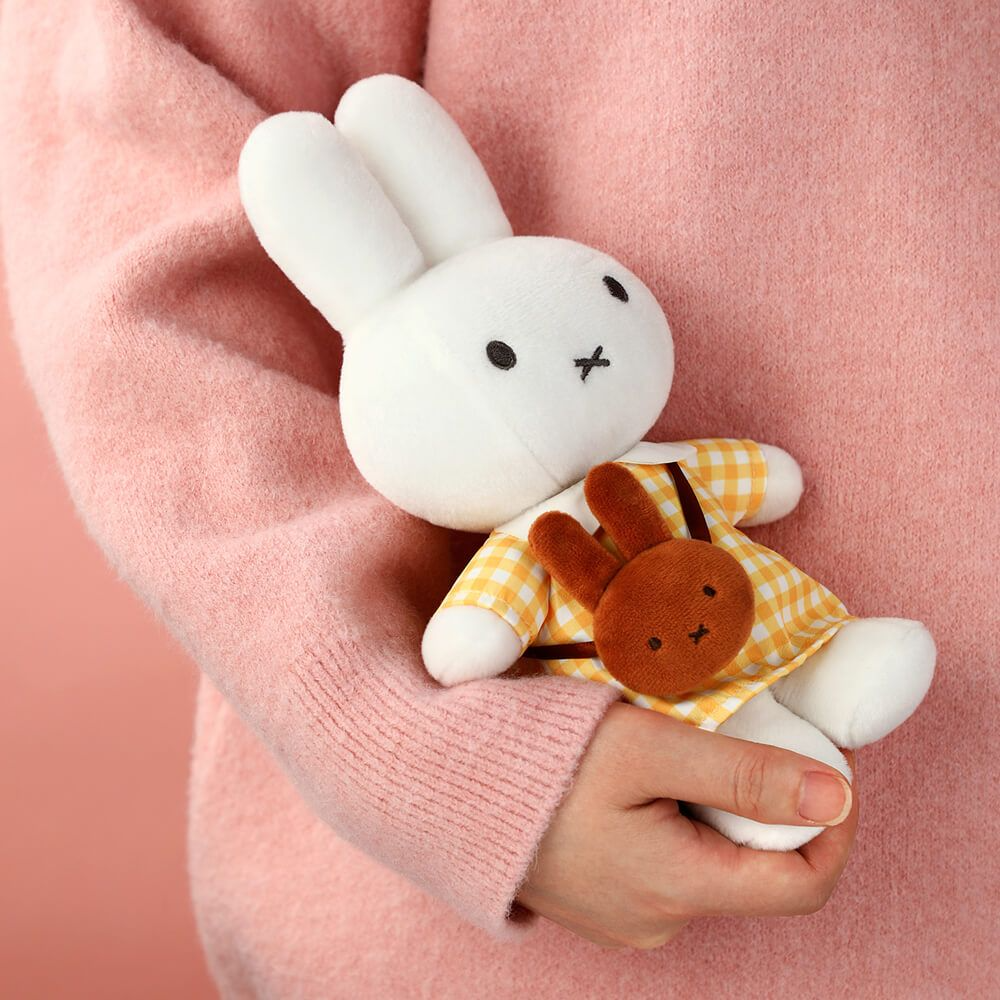 A white plush bunny doll wearing a yellow-and-white gingham dress and holding a small brown plush bunny, being held in a person's hands against a soft pink sweater background.