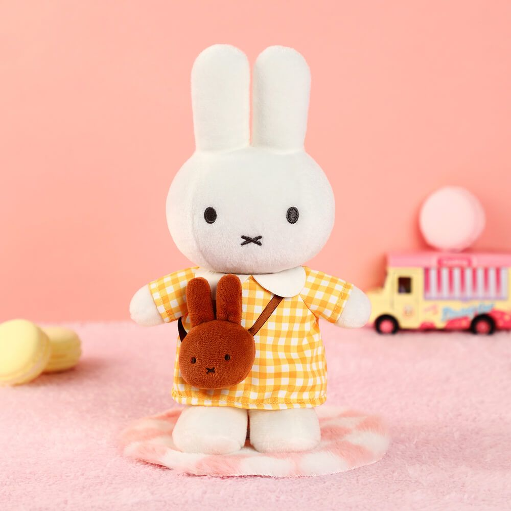 A white plush bunny doll wearing a yellow-and-white gingham dress and carrying a small brown bunny-shaped bag, posed on a pink surface against a pink background with toy props.