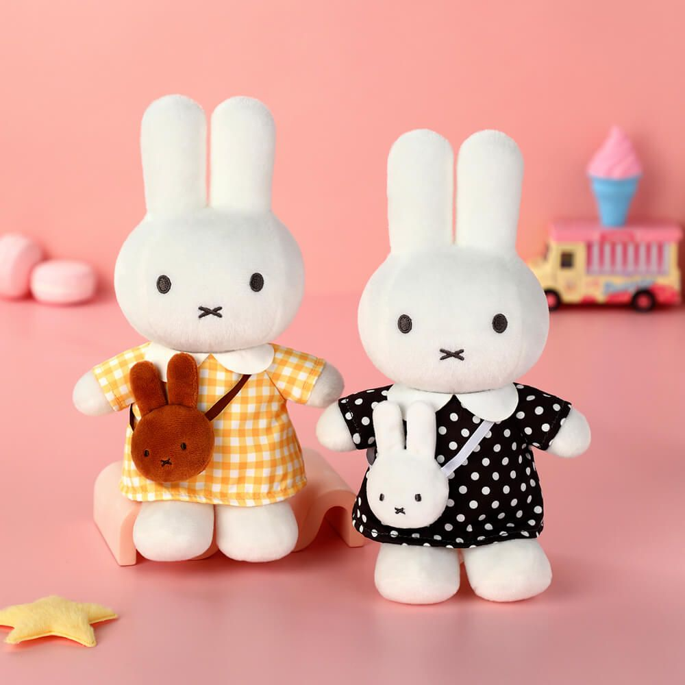 Two plush rabbit toys posed side by side on a pink surface against a pink background. The left rabbit wears a yellow-and-white gingham dress and carries a small brown bunny-shaped bag.