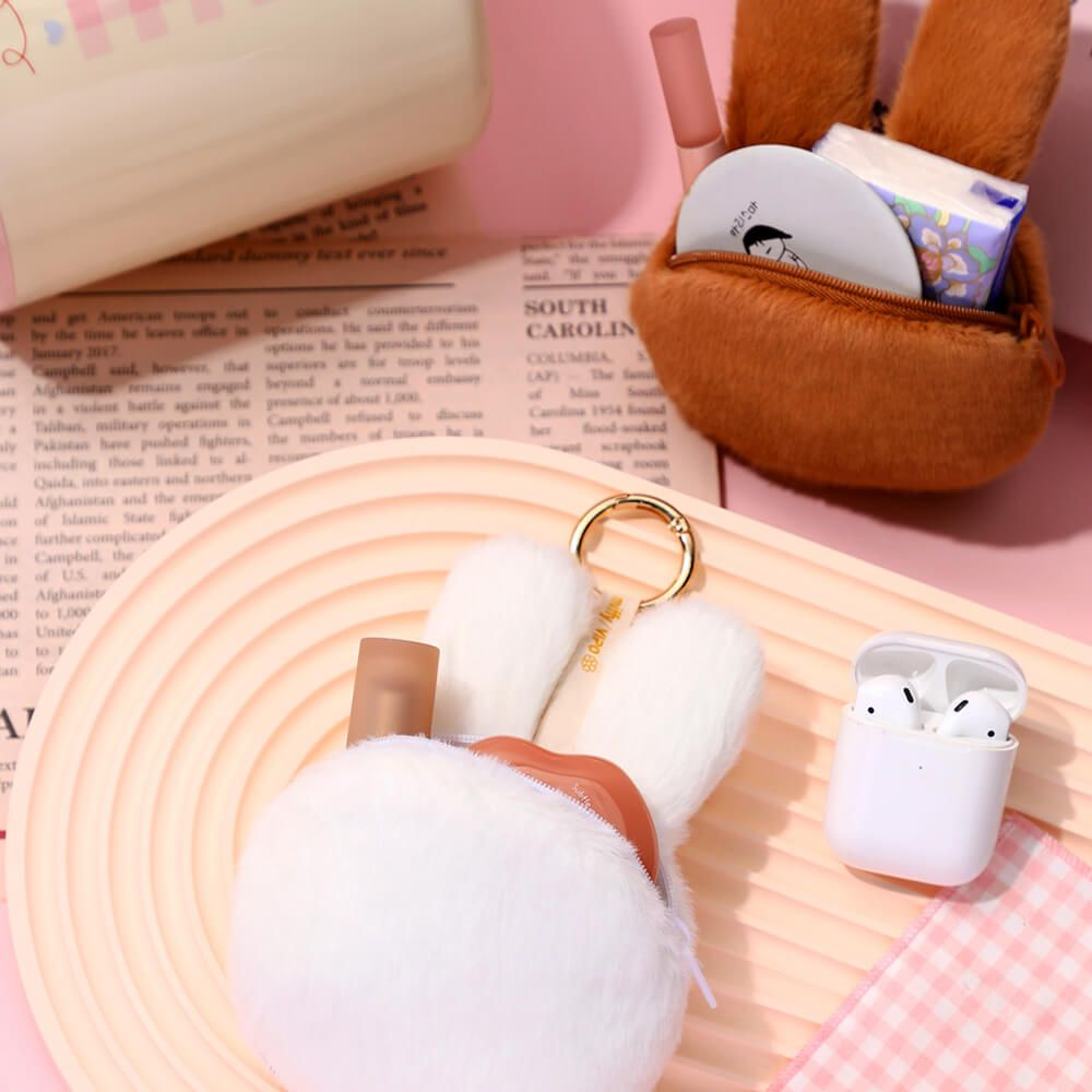 White wireless earbuds with a brown case on a pink surface with a book and plush toy.