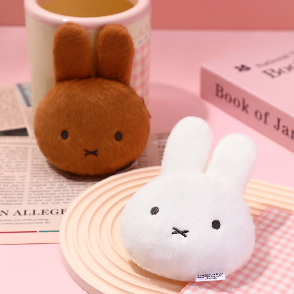 Two plush toys, one brown and one white, on a pink surface with a book and mug in the background.