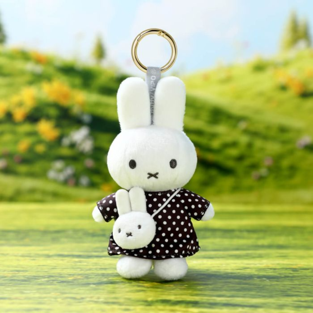 A plush Miffy keychain featuring a white bunny in a black dress with white polka dots, holding a small matching plush bag, set against a blurred green outdoor background.