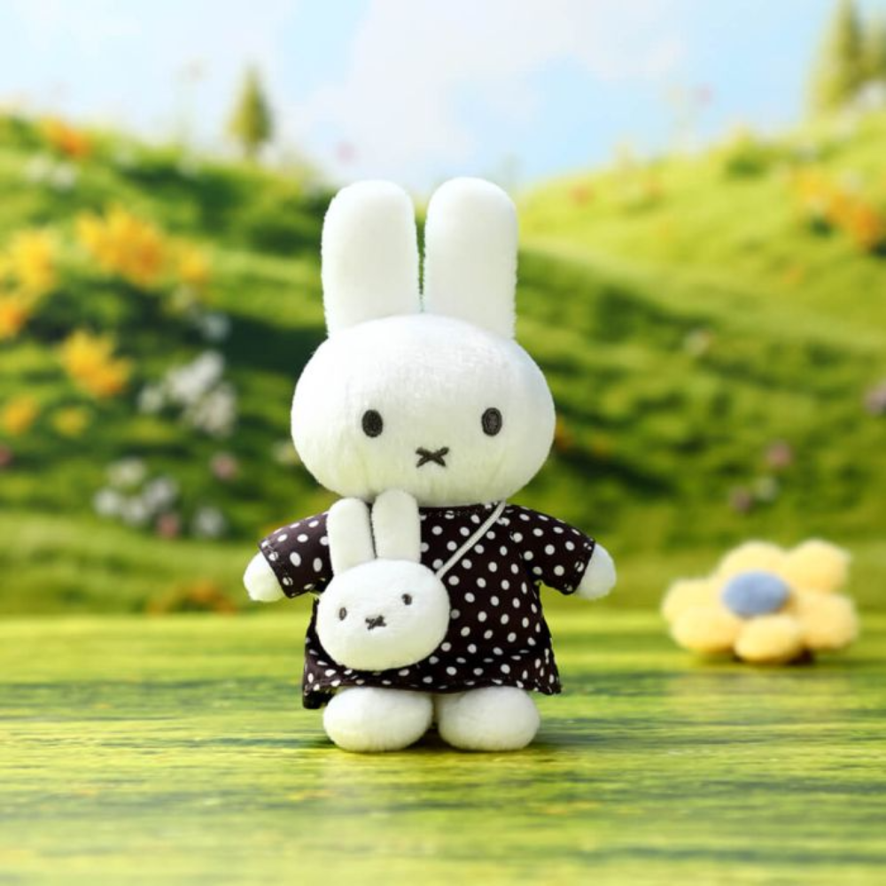 A white plush bunny character standing on a green surface, wearing a black dress with white polka dots and carrying a small matching white bunny bag. The background is a blurred outdoor-style scene with green hills, flowers, and blue sky.