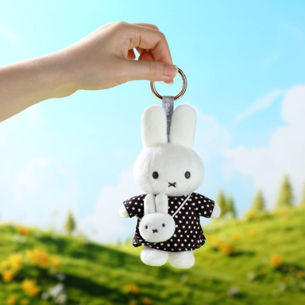 A hand holds a plush rabbit keychain with a small matching plush bag against a blurred outdoor background of green grass, yellow flowers, and a blue sky.