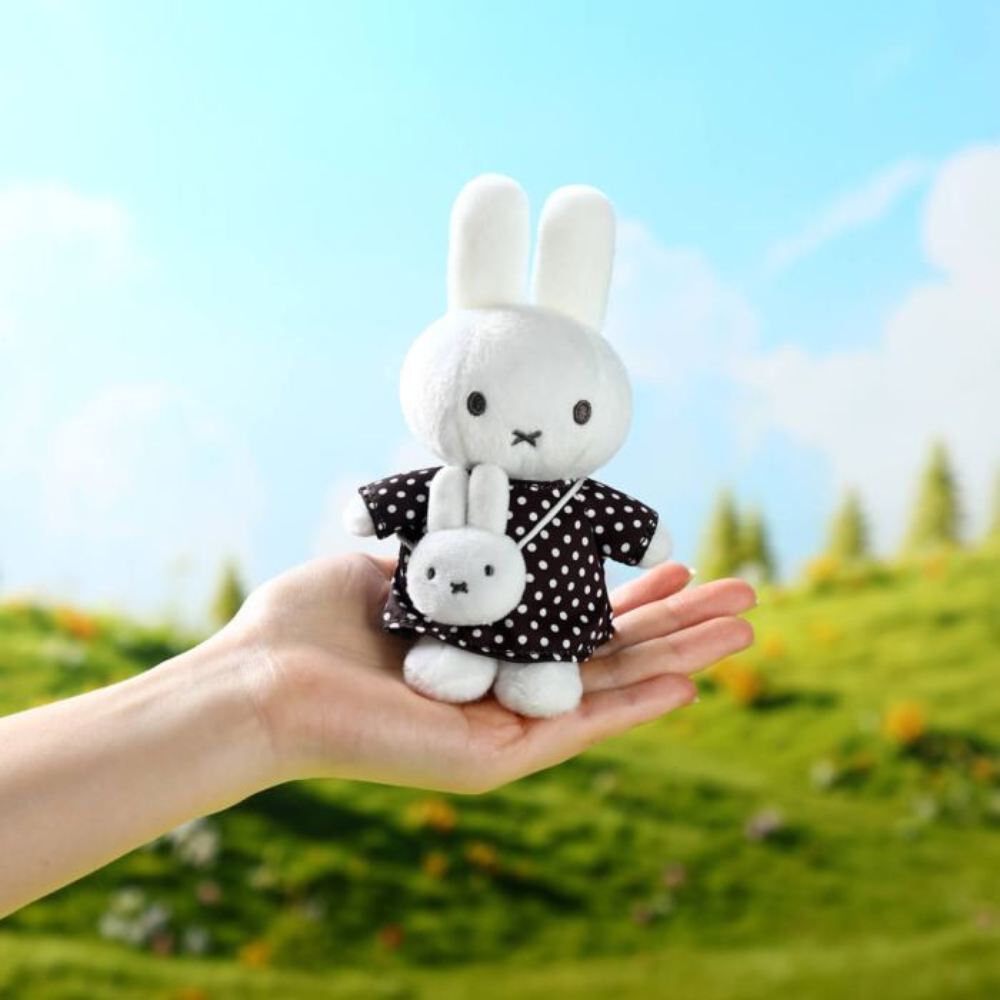 A hand holds a small white plush rabbit doll wearing a black dress with white polka dots, with a smaller matching plush rabbit in front of it, against a blurred outdoor background of green grass and blue sky.