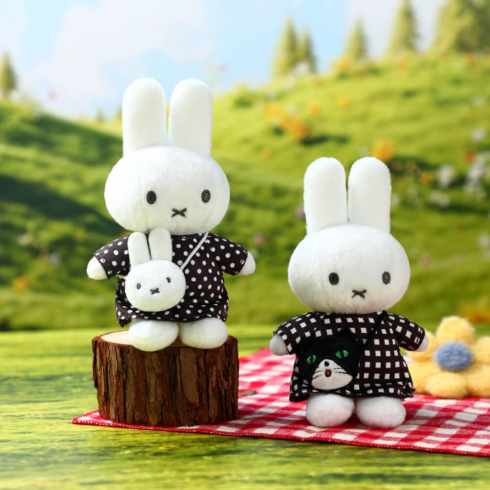 Two white rabbit plush toys dressed in black-and-white checkered outfits, posed on a red-and-white checkered blanket with a blurred green hillside background.