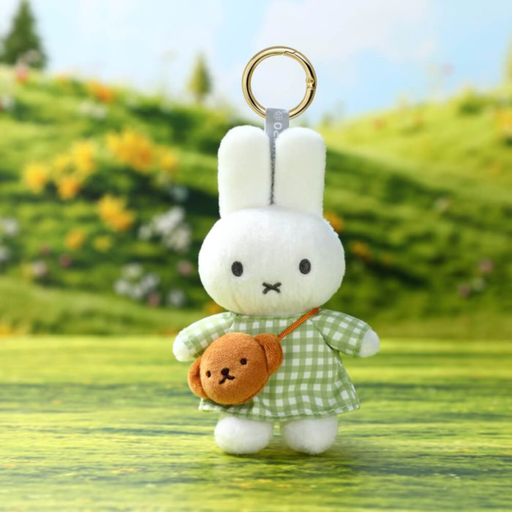 A plush keychain of a white bunny wearing a green and white checkered dress and carrying a small brown crossbody bag, set against a blurred outdoor meadow background.