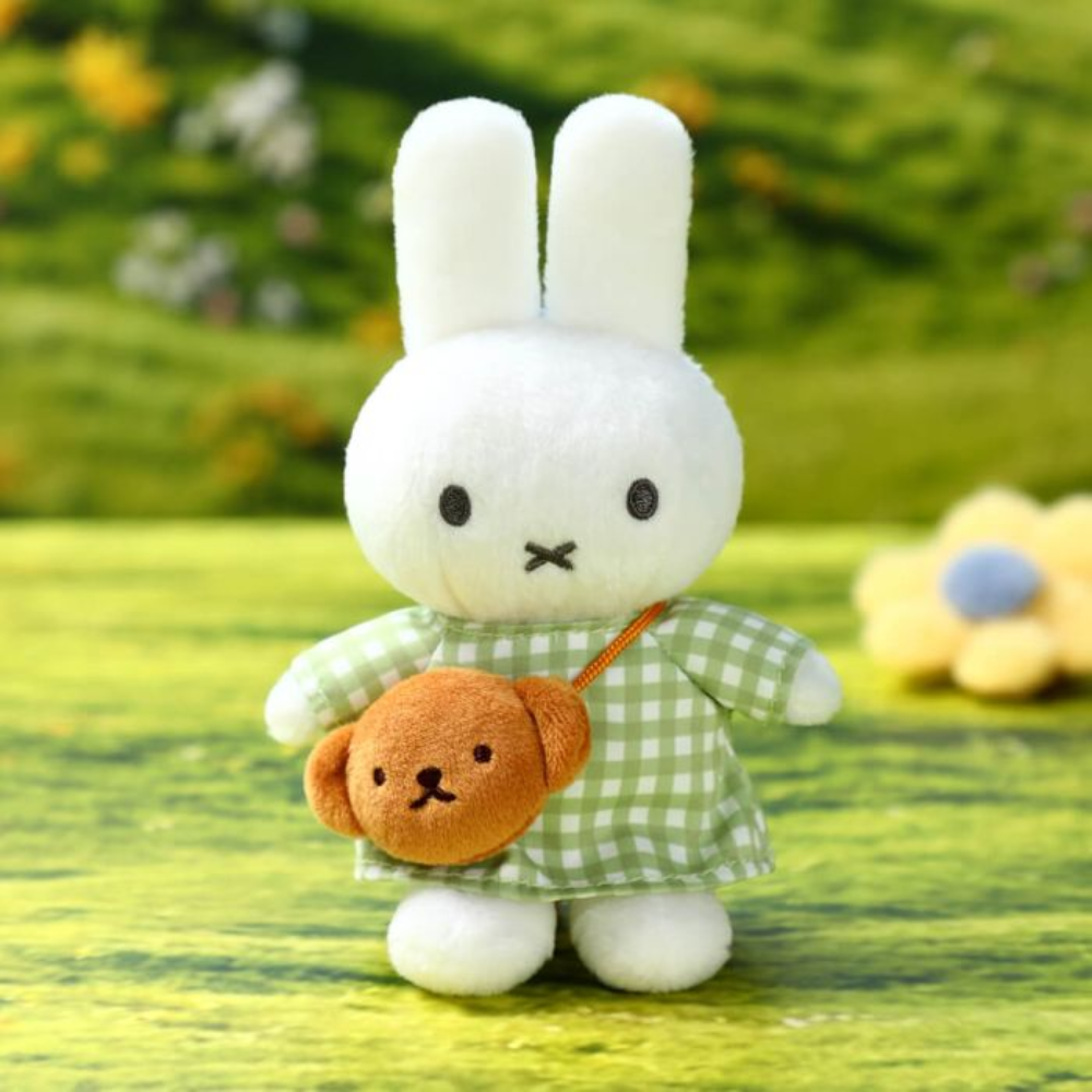 A white plush bunny doll wearing a green-and-white check dress and carrying a small brown plush bag, standing on a blurred green outdoor-style background.