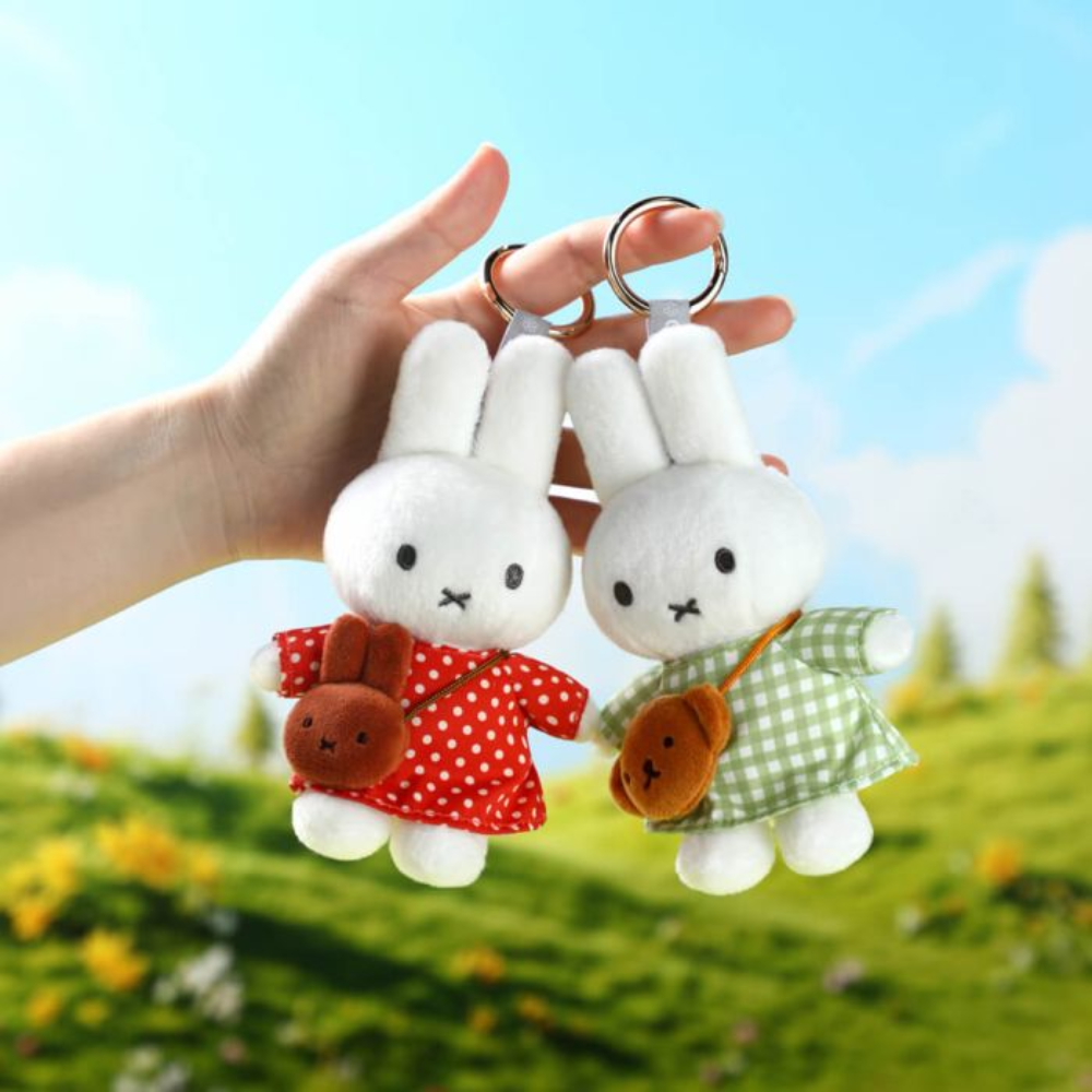 Two plush white bunny keychains held in a person's hand against a blurred outdoor background. One bunny wears a red dress with white polka dots and carries a brown bag, while the other wears a green gingham dress and carries a brown bag.