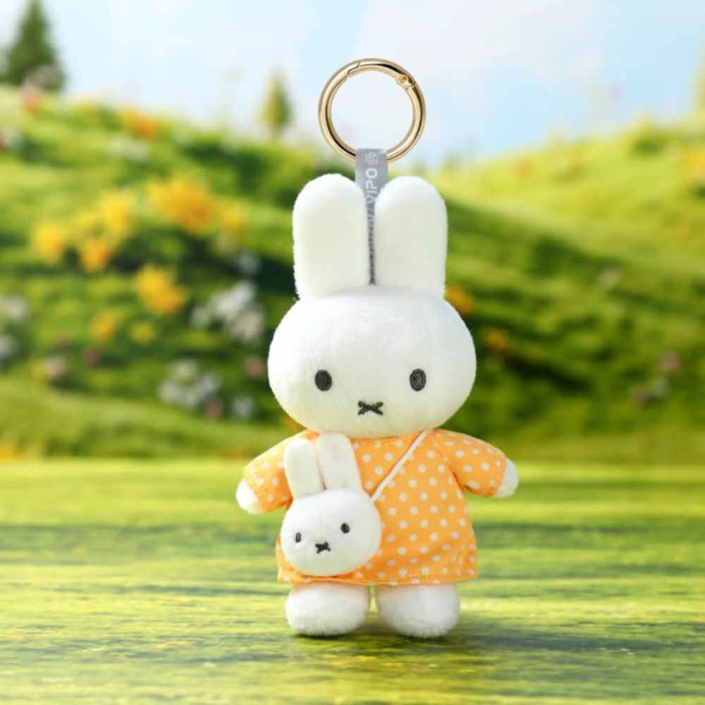 A plush keychain featuring a white bunny character wearing an orange dress with white polka dots and holding a smaller matching plush, attached to a gold key ring. The background is a blurred outdoor-style green landscape.