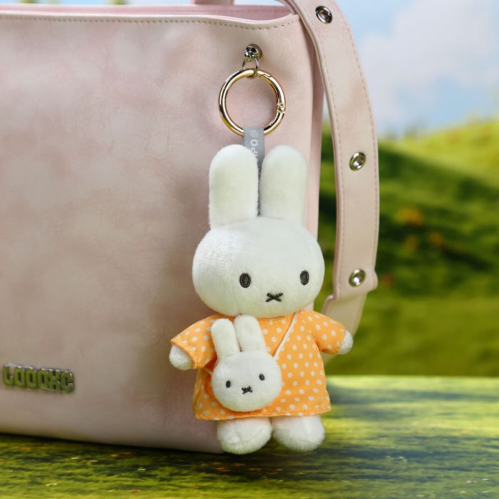 A plush rabbit keychain charm with long white ears, wearing an orange dress with white polka dots, hanging from a gold ring on a pale pink handbag. The background is a softly blurred outdoor scene with green grass and blue sky.
