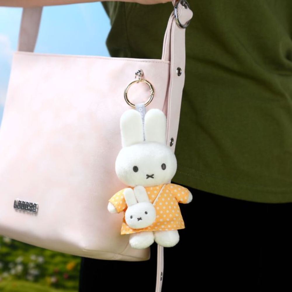 A pale pink shoulder bag with a plush white bunny keychain accessory wearing an orange polka-dot dress, shown being carried outdoors by a person in an olive green top.