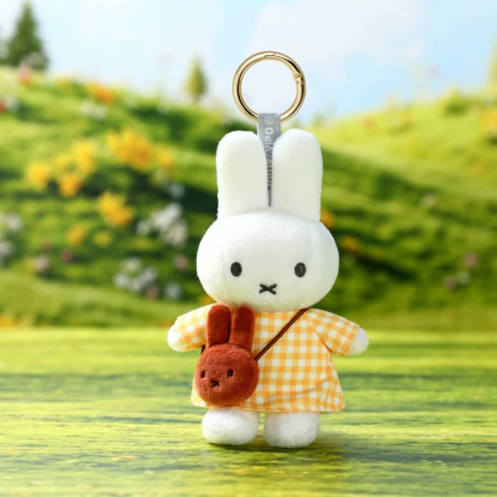 A plush keychain of Miffy the white rabbit wearing an orange and yellow gingham dress, holding a small brown crossbody bag, with a gold metal key ring at the top. The background is a soft-focus outdoor scene of a green hill with flowers.