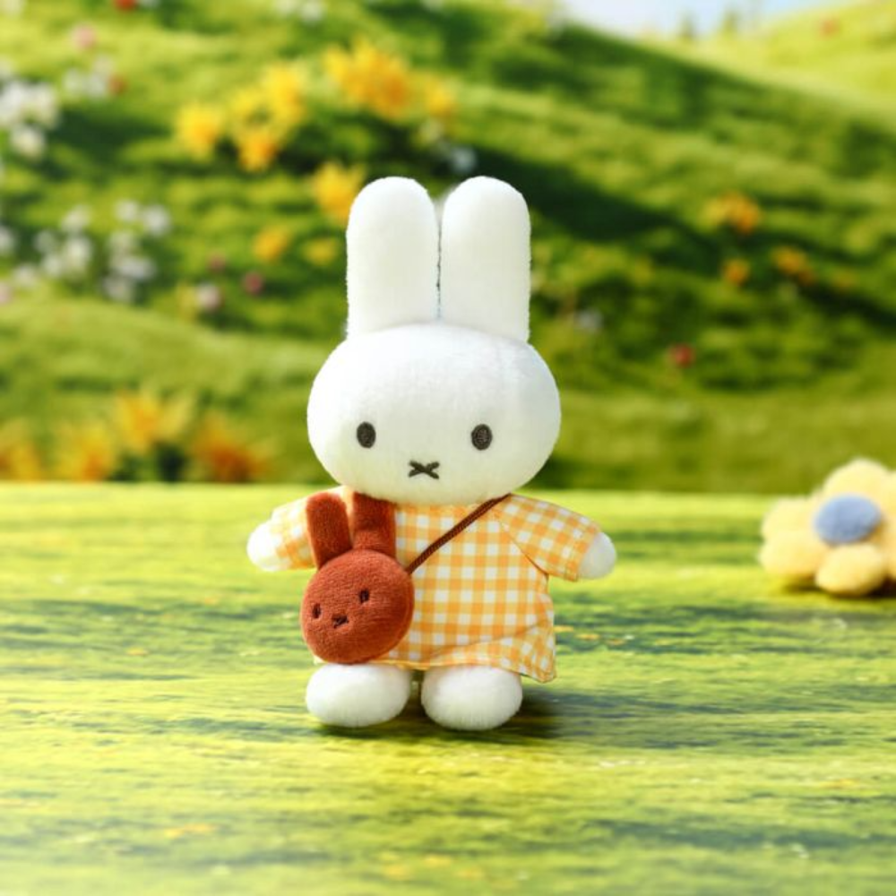 A small plush rabbit toy standing on a grassy surface, wearing an orange-and-white gingham dress and carrying a brown crossbody bag with a rabbit face. The background is a blurred green outdoor hillside with flowers.