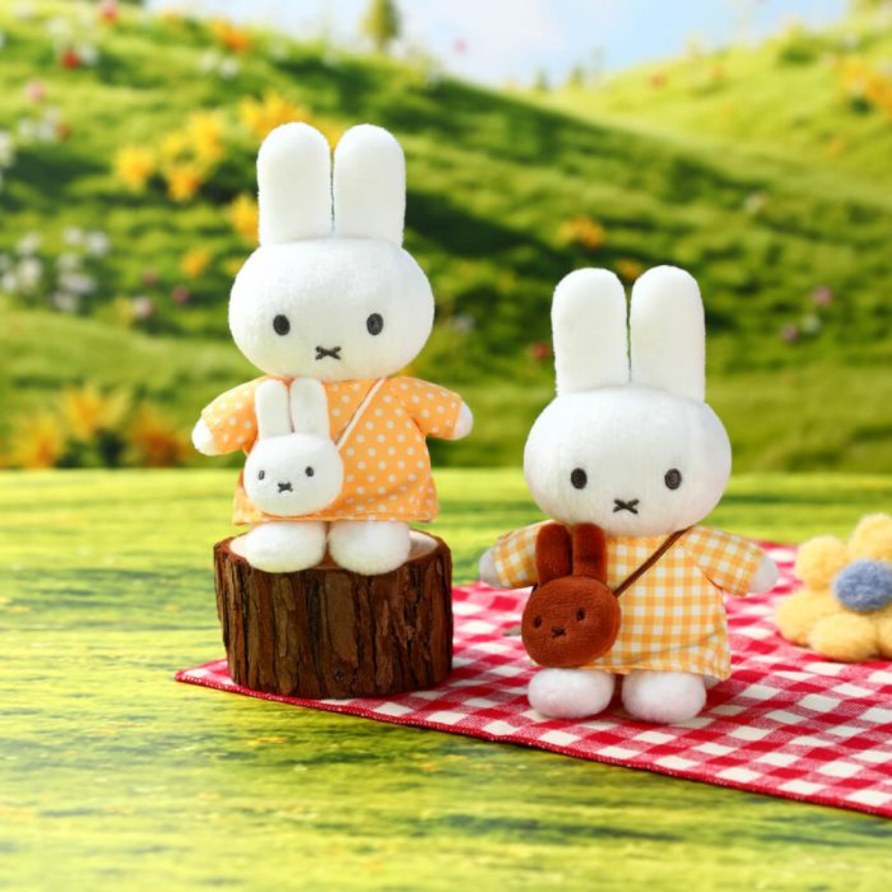 Two plush rabbit toys dressed in orange and yellow polka-dot outfits, posed on a red-and-white checkered blanket with a blurred grassy hill background.