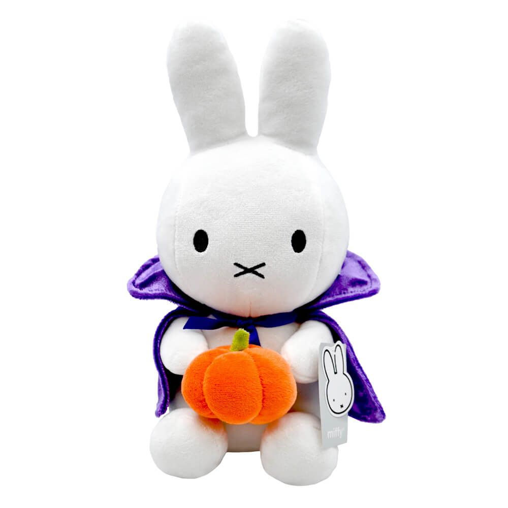 A white plush rabbit toy with long ears, dressed in a purple cape and holding a small orange pumpkin, shown on a plain white background. A hangtag with the Miffy bunny logo is attached to the side.