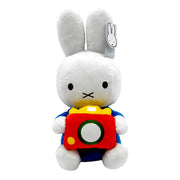A white plush rabbit toy holding a red camera with yellow, green, and white details, wearing a blue outfit. A small hangtag is attached to one ear.