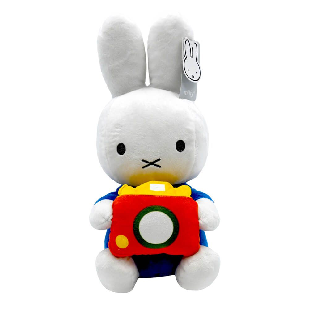 A white plush rabbit toy holding a red camera with yellow, green, and white details, wearing a blue outfit. A small hangtag is attached to one ear.
