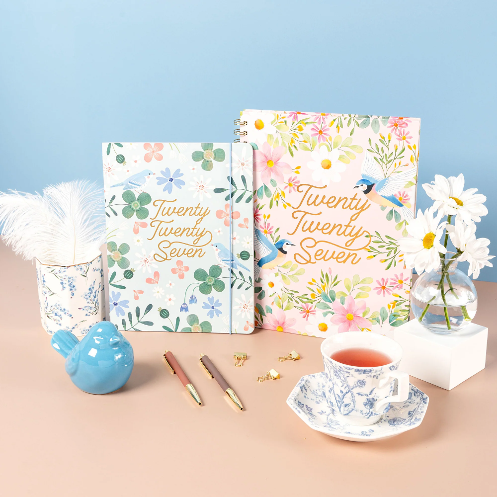 Two floral notebooks with gold foil 'Twenty Seven' text are displayed on a peach surface against a light blue background, styled with a blue ceramic bird, pens, a teacup, and white daisies.
