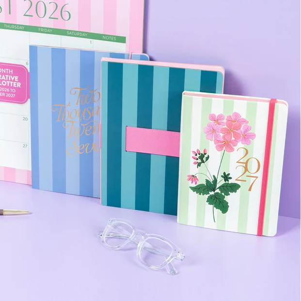 A coordinated stationery set arranged on a lavender surface against a matching lavender background, including a large August 2026 desk calendar, striped planner/notebooks, a floral 2027 notebook, a clear desk organizer, a pen, and clear eyeglasses.