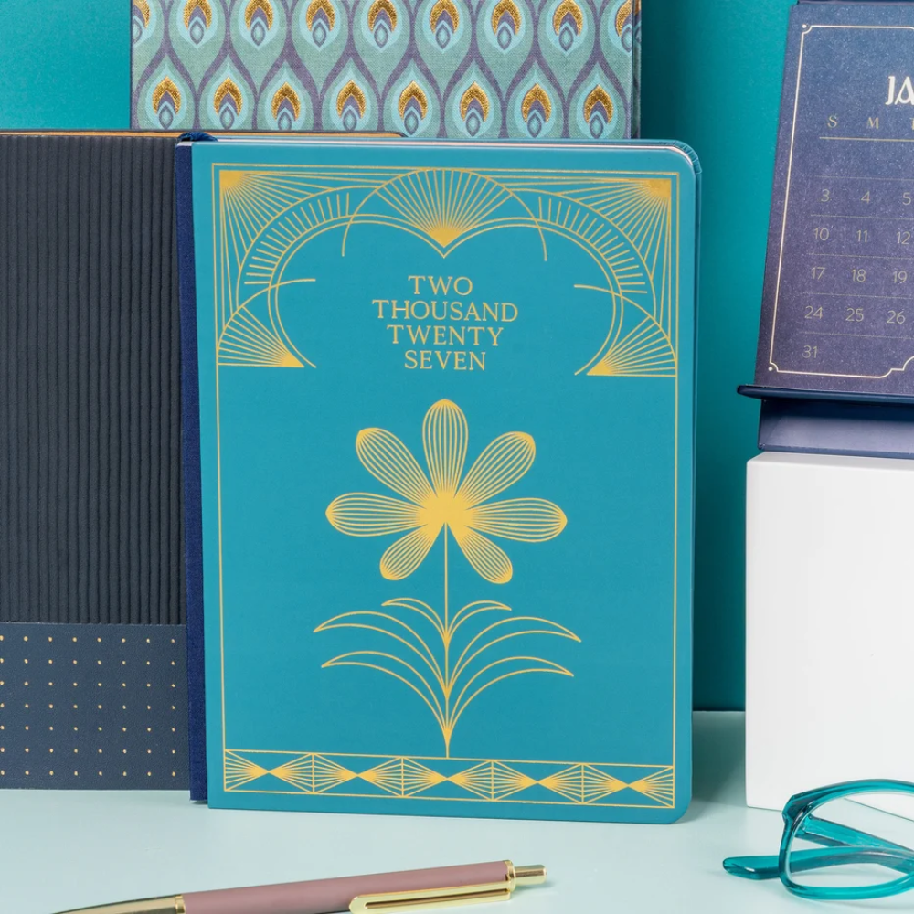 A teal and gold hardcover notebook or journal standing upright on a tabletop, surrounded by desk props including patterned paper, a calendar, eyeglasses, and a pen.