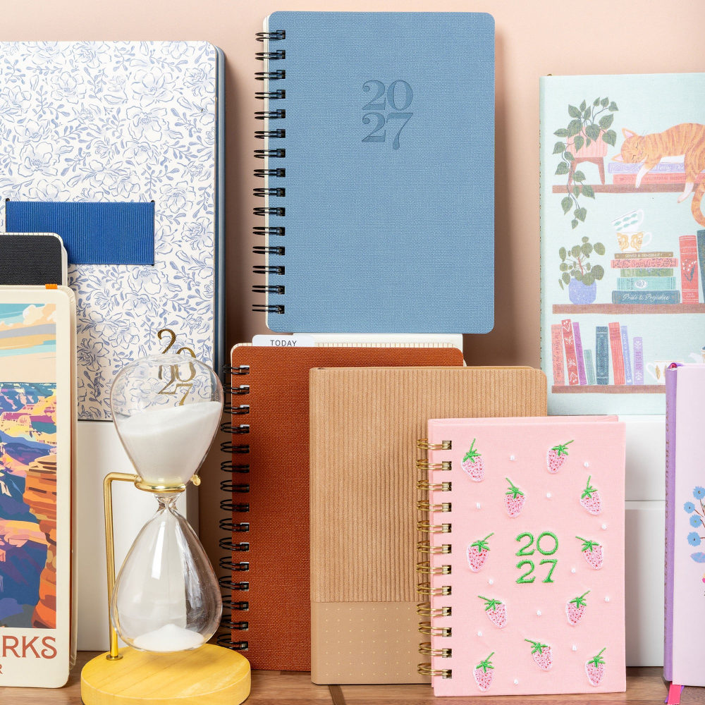 A collection of spiral-bound notebooks and planners arranged on a shelf against a soft peach background. Visible items include a blue 2027 planner, a pink strawberry-themed 2027 notebook, a brown kraft notebook, a white patterned planner, and other stationery with colorful illustrations.
