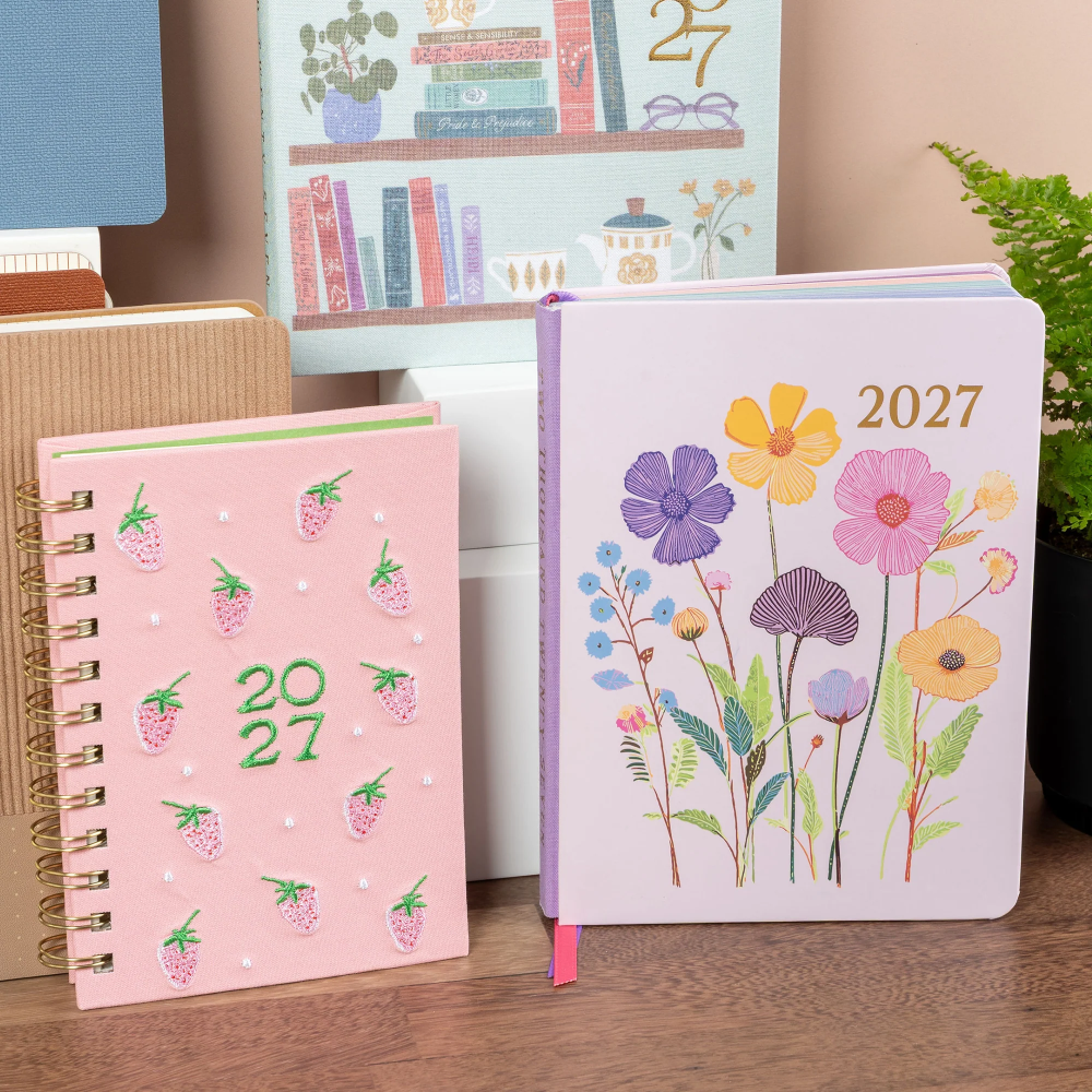 Two decorative 2027 planners on a wooden desk, one pink spiral-bound with strawberry illustrations and the other a lavender hardcover with colorful wildflower illustrations. A bookshelf and plant are visible in the softly lit background.