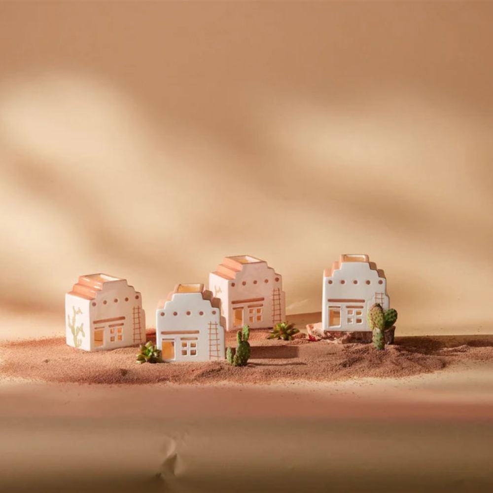 Four miniature white and peach-colored house models arranged on a small sandy platform with tiny green cacti, set against a warm beige background with soft shadow overlays.