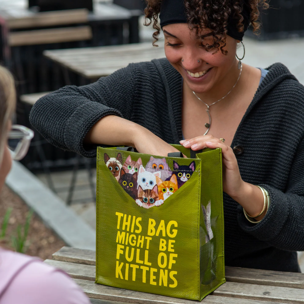 Person holding a green bag with cat illustrations and text, sitting at an outdoor table.