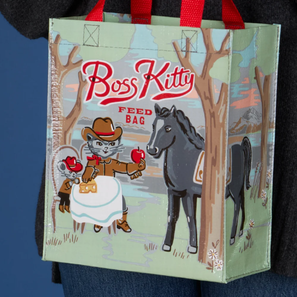 A person in a dark gray sweater holds a Blue Q / Boss Kitty reusable shopping bag with red handles. The bag has a light green background with an illustration of a cat and a horse in a forest.