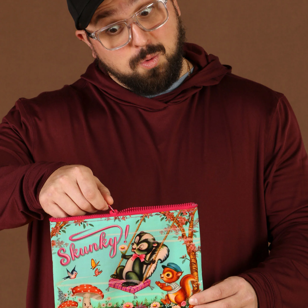 A man wearing a black cap, clear glasses, and a burgundy hoodie holds up a colorful zippered pouch with a cartoon raccoon illustration and the text 'Skunky!'.