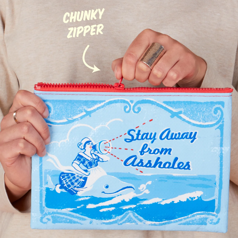 A person holds a light blue zipper pouch with a retro illustration of a woman flying a kite and the text 'Stay Away from Assholes.' The person's hands are visible, along with a red zipper and a hand-written label pointing to the zipper with the words 'CHUNKY ZIPPER.