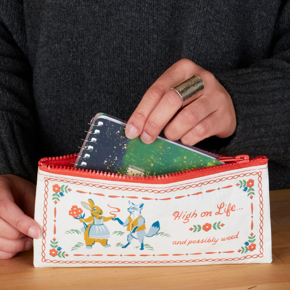 A person holds an open white zip pouch with a red zipper and a colorful illustrated print on the front, showing it being used to store art supplies. The pouch is placed on a wooden table, with text at the bottom reading 'Perfect for carrying art supplies, makeup brushes and more'.