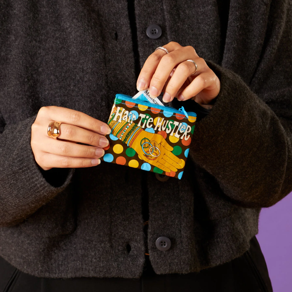 A person wearing a dark charcoal knit cardigan holds a small colorful coin purse labeled 'HATE THE MISTLE' against a solid purple background.