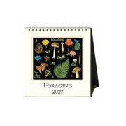 A spiral-bound 2027 desk calendar titled 'FORAGING' with a cream-colored cover, featuring a black square illustration panel filled with colorful botanical and mycological drawings, and a dark side panel forming the stand.