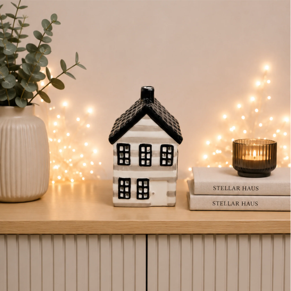 A small ceramic house-shaped candle holder or decor piece with a black roof and black window details on a white body, displayed on a wooden surface beside books, a candle, and a plant in a vase, with warm string lights in the background.