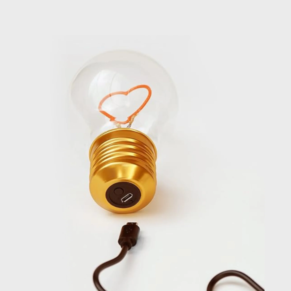 A clear glass light bulb with a gold screw base and an orange heart-shaped filament, shown on a white background with a dark brown power cord.