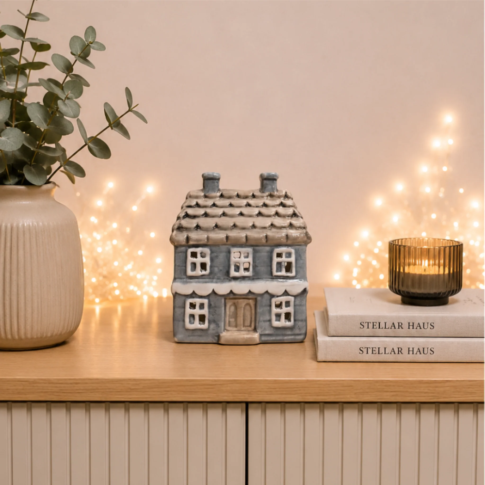 A small gray ceramic house-shaped container or decor piece sits on a light wood cabinet beside a beige vase with eucalyptus, a stack of Stellar Haus books, and a lit amber candle, with warm fairy lights blurred in the background.