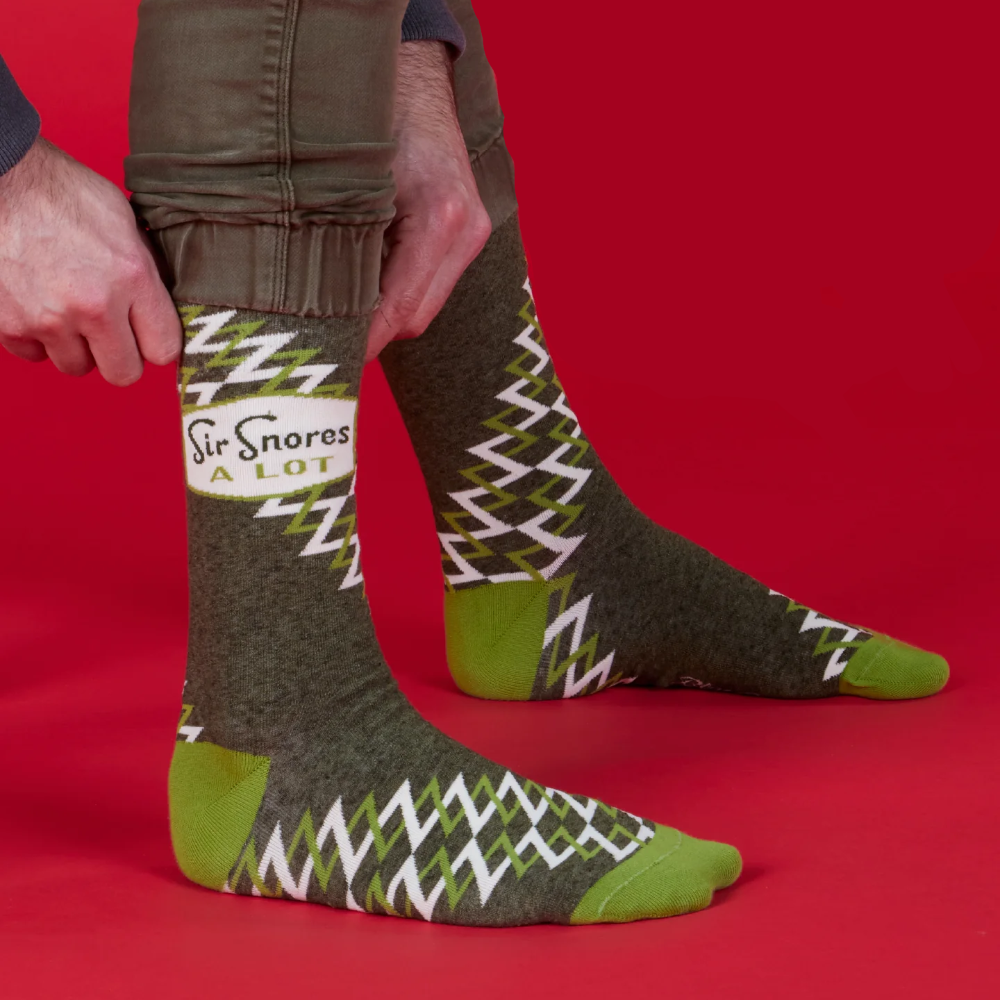 A person wearing olive green socks with a white and green zigzag pattern and bright green toes and heels, shown against a solid red background with the text 'Nylon for strength'.