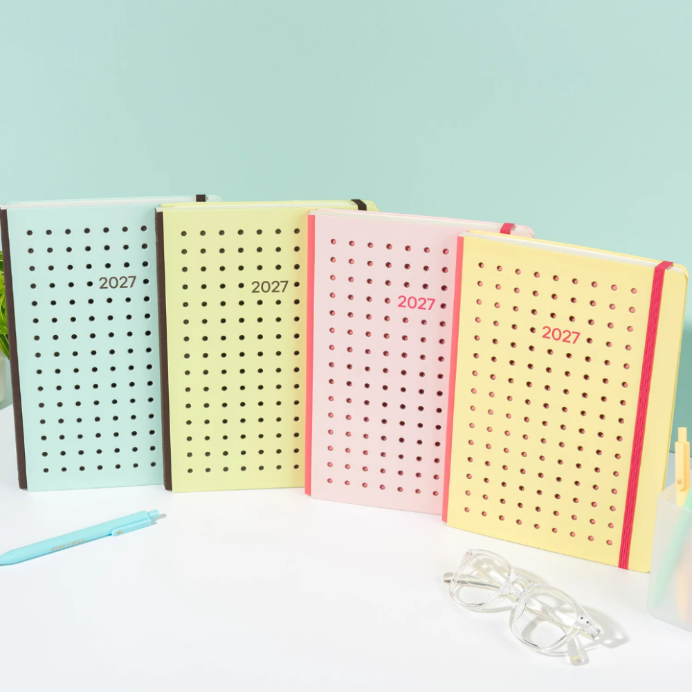 Four pastel-colored 2027 daily planners with perforated dot covers are lined up on a white surface. The planners are in light blue, pale yellow, pink, and pale yellow, each with an elastic closure and the year '2027' printed on the front.