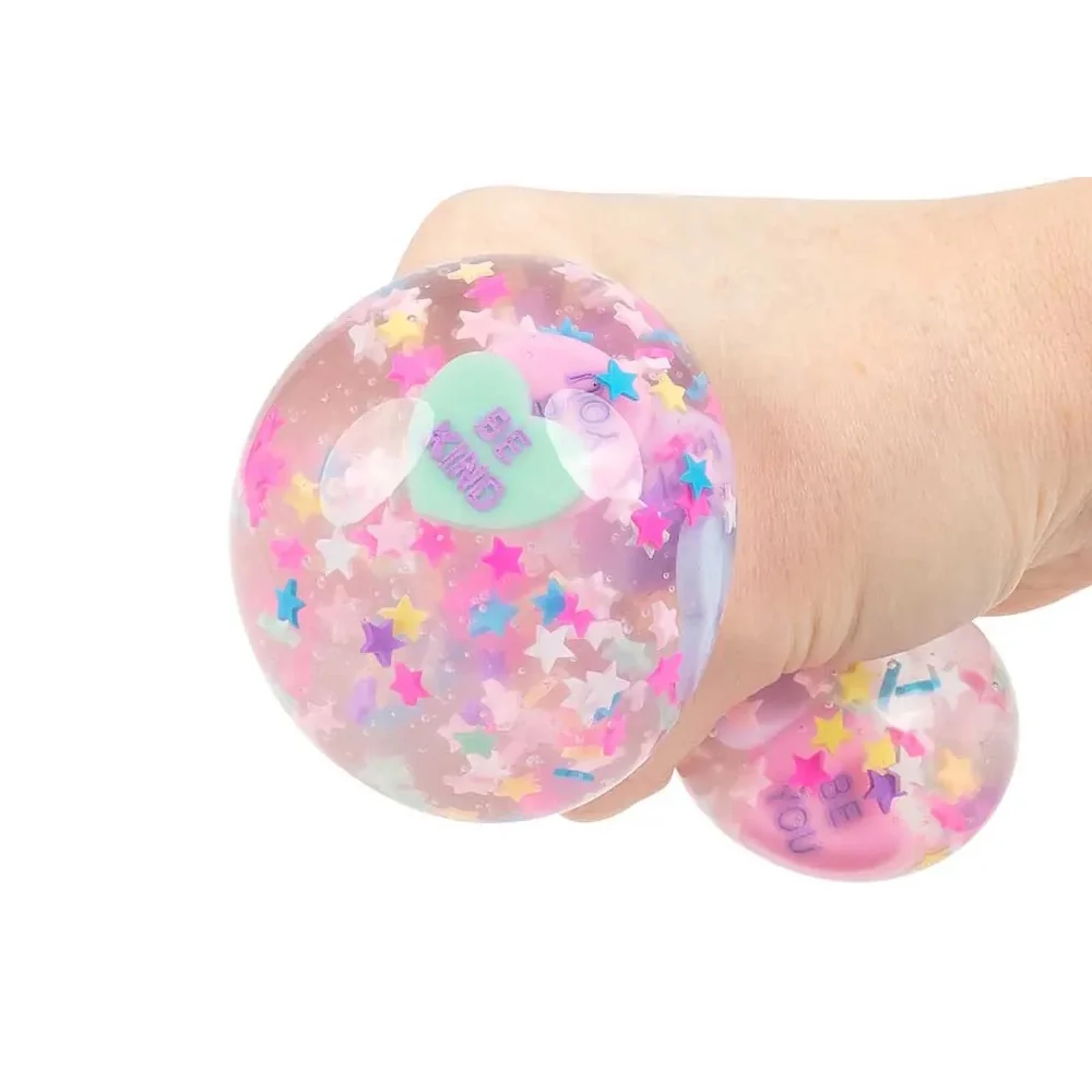 A hand squeezing a clear stress ball toy filled with pastel pink glitter and multicolored star confetti, with a mint green heart-shaped tag that reads 'BEST WISHES'.