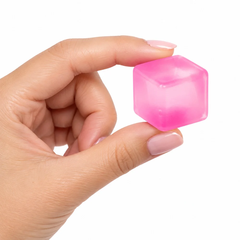A hand with light pink manicured nails holds a small, translucent pink cube-shaped object against a solid white background.