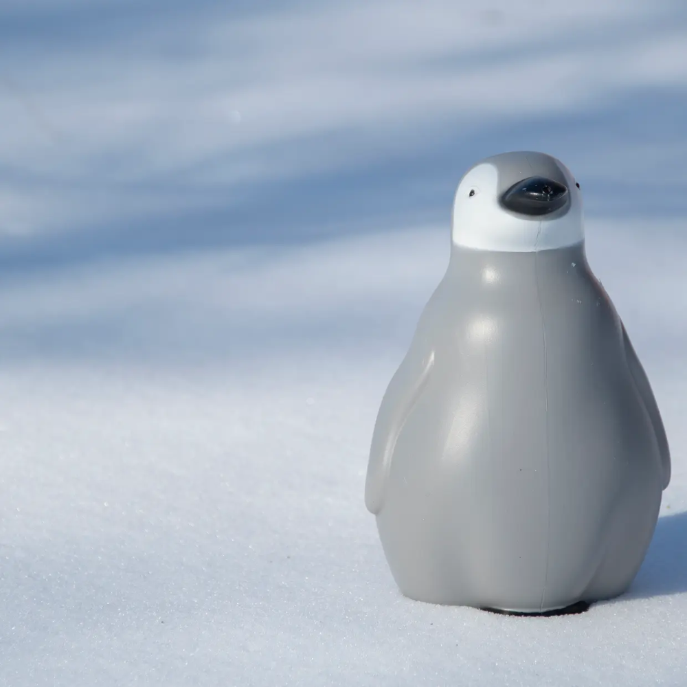 A small penguin-shaped product, possibly a toy or decorative figure, standing upright in the snow against a soft blue sky background.