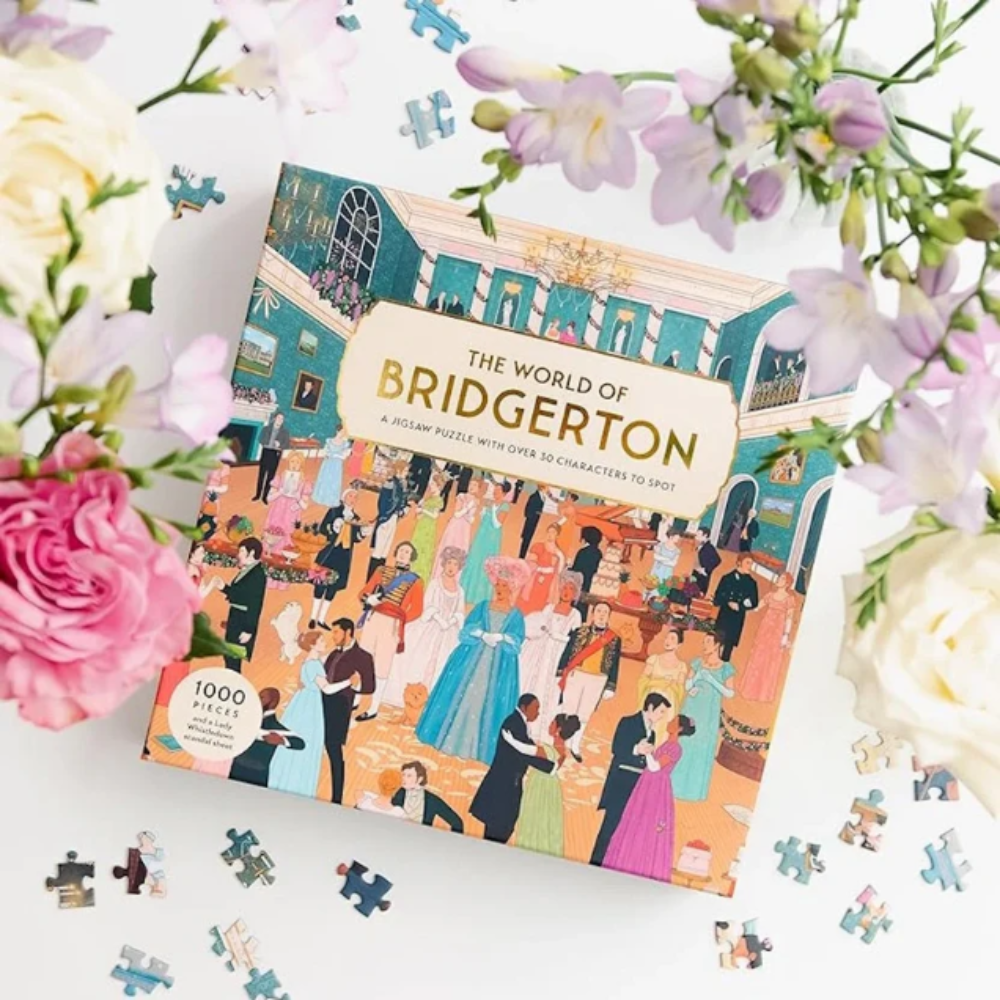 A 1000-piece jigsaw puzzle box titled 'The World of Bridgerton' is displayed on a white surface surrounded by pink and white flowers, with a few loose puzzle pieces scattered around it.