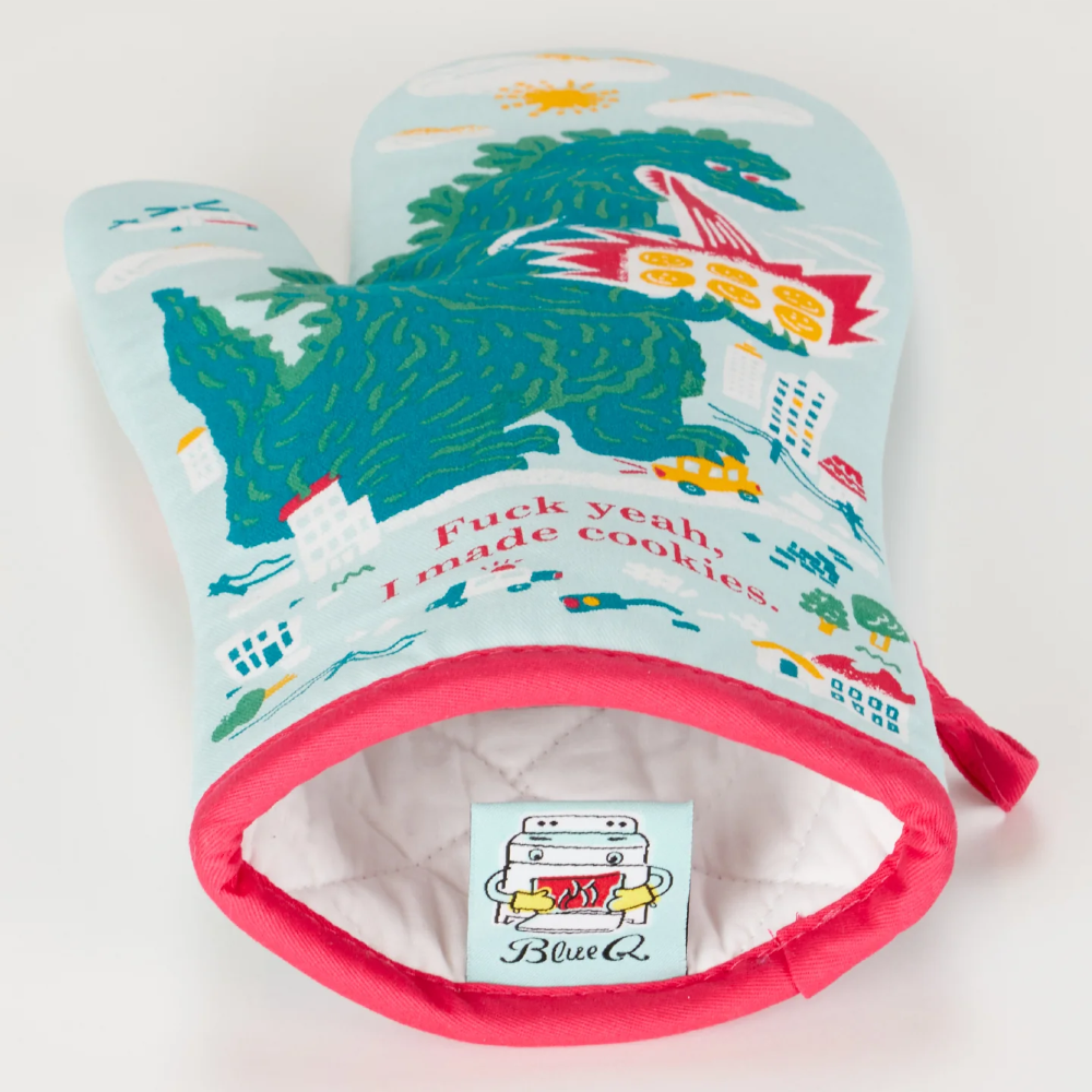 A novelty oven mitt with a light blue background and a colorful cartoon dinosaur scene, featuring the text 'Fuck yeah, I made cookies.' The mitt has bright pink trim and a white quilted interior with a visible 'Blink6' brand patch.